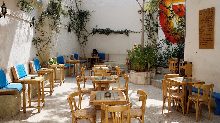 Sunlit courtyard at Jadal for Knowledge and Culture in Jabal Al Weibdeh, Amman, with wooden tables, blue cushioned benches, climbing plants and a mural