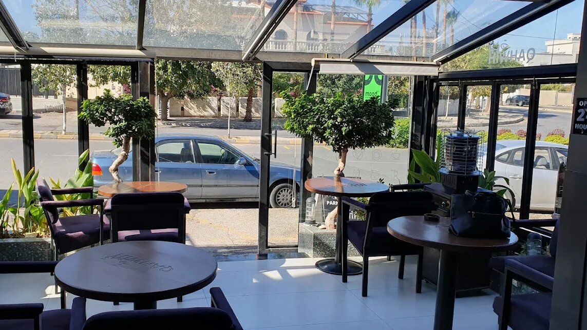 Glass-walled conservatory seating at Qahwih cafe in Abdoun, Amman, with branded round tables, padded armchairs, plants and daylight