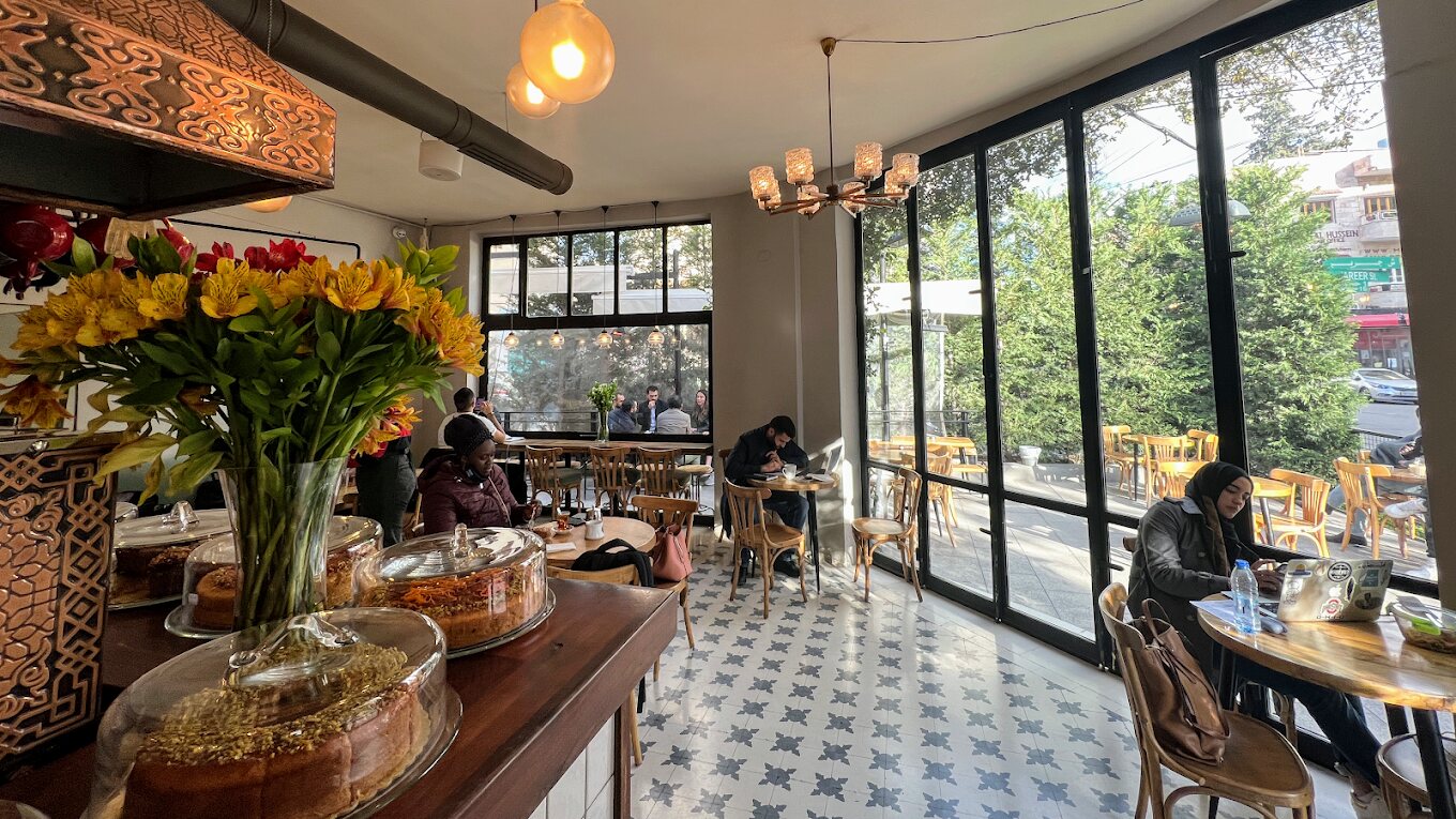 Interior of Rumi Cafe in Jabal Al Weibdeh, Amman, with patterned tiled floor, bentwood chairs, a cake counter, floor-to-ceiling windows and customers working on laptops