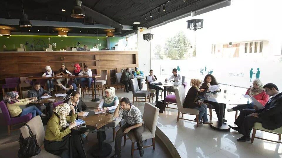 Students and professionals working at tables in the open-plan co-working area at V Business Center, Amman, with a glass-walled meeting room